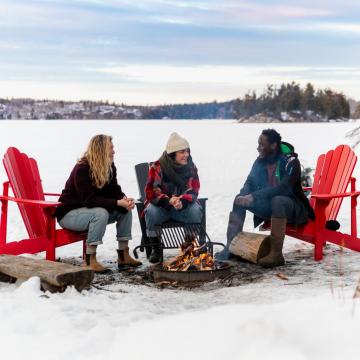 In einer Schneelandschaft am Falcon Lake in Manitoba sitzen drei junge Menschen am Lagerfeuer.