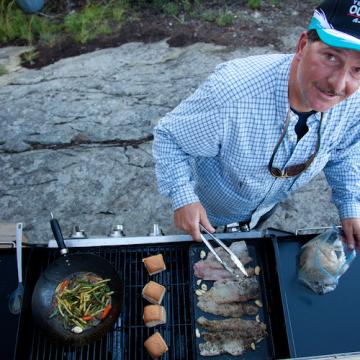 Ein Mann steht an einem Grill und bereitet am Seeufer fangfrischen Fisch zu.