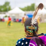 Eine indigene Frau mit traditioneller Tracht steht mit dem Rücken zur Kamera und blickt auf eine Zeremonie-Szene.