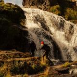 Ein Wanderer spring über einen Stein vor einem Wasserfall