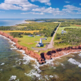 Blick auf Prince Edward Island aus der Luft, zu sehen ist die rote schroffe Küste, ein Leuchtturm und weites grünes Land