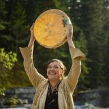 Eine Indigene hebt eine Trommel in die Höhe, präsentiert sie mit einem Lächeln vor einer Waldlandschaft